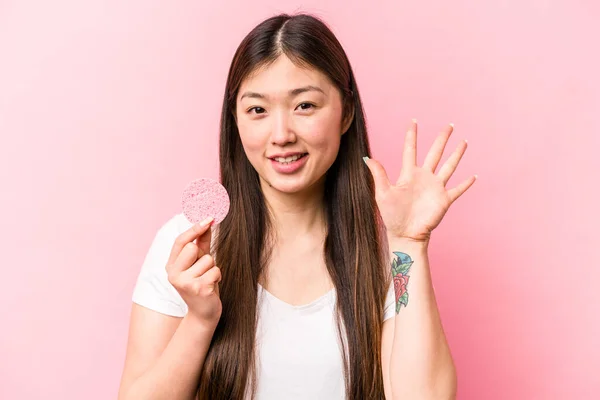 年轻的亚洲女人 手握粉红背景的面部海绵 笑容满面 露出五号手指 — 图库照片