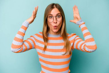 Mavi arka planda izole genç beyaz kadın zaferi ya da başarıyı kutluyor, şaşırıyor ve şok oluyor..