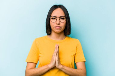 Genç İspanyol kadın mavi arka planda izole edilmiş dua ediyor, bağlılık gösteriyor, dindar biri ilahi ilham arıyor..