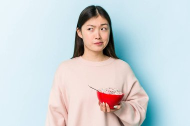 Mavi arka planda bir kase mısır gevreği tutan genç Asyalı kadının kafası karışmış ve kendinden emin değil..