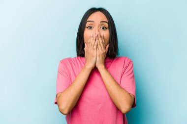Mavi arka planda izole edilmiş genç İspanyol kadın elleriyle ağzını kapatırken şok oldu..