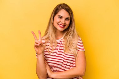 Sarı arka planda izole edilmiş genç beyaz kadın neşeli ve kaygısız parmaklarıyla barış sembolü gösteriyor..