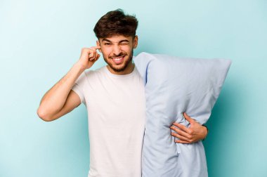 Pijama giyen genç İspanyol bir adam yastık tutuyor mavi arka planda izole edilmiş kulakları elleriyle kapatıyor..