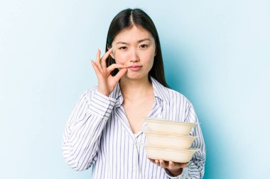 Saklama kabı taşıyan Asyalı genç bir kadın mavi arka planda izole edilmiş dudaklarında parmakları olan bir sır saklıyor..