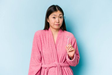 Mavi arka planda izole edilmiş jilet tutan genç Asyalı kadın omuzlarını silker ve gözleri açık kalır..