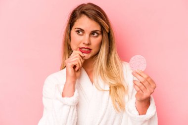 Elinde yüz süngeri tutan beyaz bir kadın pembe arka planda izole edilmiş. Bir şeyin fotokopi odasına baktığını düşünüyor..