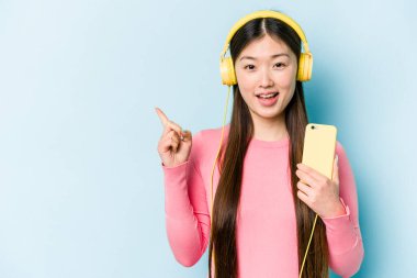 Genç Asyalı bir kadın mavi arka planda izole edilmiş müzik dinliyor gülümsüyor ve boş alanda bir şeyler gösteriyor..