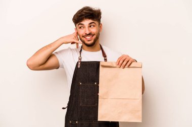 Beyaz arka planda izole edilmiş bir çanta taşıyan genç İspanyol tezgahtar parmaklarıyla bir cep telefonu hareketi gösteriyor..