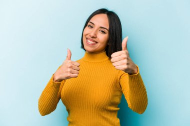 Genç İspanyol kadın mavi arka planda izole edilmiş. Baş parmağını kaldırıyor, gülümsüyor ve kendinden emin..
