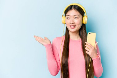 Genç Asyalı bir kadın mavi arka planda izole edilmiş müzik dinlerken bir avuç içinde fotokopi alanı ve belinde başka bir el tutarken görülüyor..