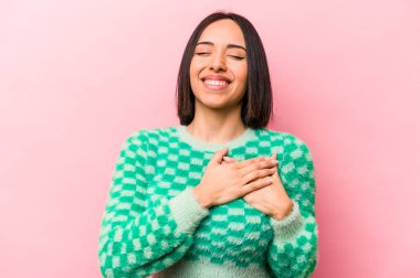 Pembe arka planda izole edilmiş genç İspanyol kadın gülüyor, ellerini kalbe koyuyor, mutluluk kavramını koruyor..