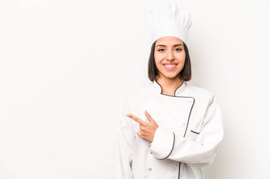 Beyaz arka planda izole edilmiş genç İspanyol aşçı kadın gülümsüyor ve boş alanda bir şeyler gösteriyor..