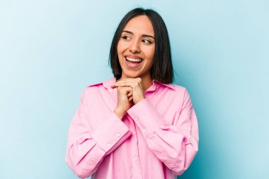 Mavi arka planda izole edilmiş genç İspanyol kadın ellerini çenesinin altında tutuyor ve mutlu bir şekilde yan tarafa bakıyor..