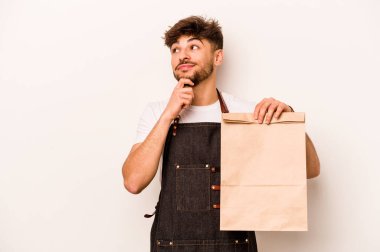 Beyaz arka plana izole edilmiş, şüpheli ve şüpheci bir ifadeyle yan bakan bir paket taşıyan genç İspanyol tezgahtar..