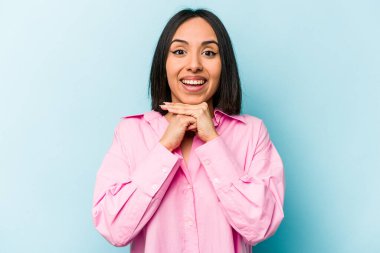 Genç İspanyol kadın mavi arka planda izole edilmiş şans için dua ediyor, şaşırmış ve ağzını açıp önüne bakıyor..