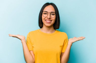 Mavi arka planda izole edilmiş genç İspanyol kadın kollarıyla ölçeği yapıyor, mutlu ve kendinden emin hissediyor..