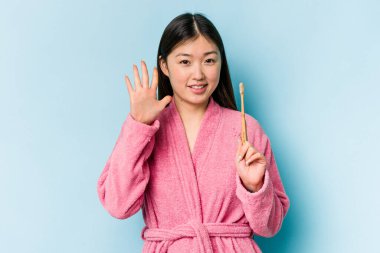 Bornoz giymiş genç Asyalı bir kadın pembe arka planda izole edilmiş dişlerini fırçalıyor gülümseyerek beş numarayı parmaklarıyla gösteriyor..
