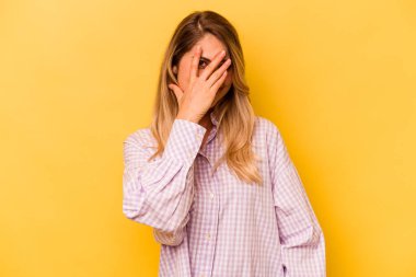 Sarı arka planda izole edilmiş genç beyaz kadın kameraya parmaklarıyla göz kırpıyor, yüzünü kapatmaktan utanıyor..