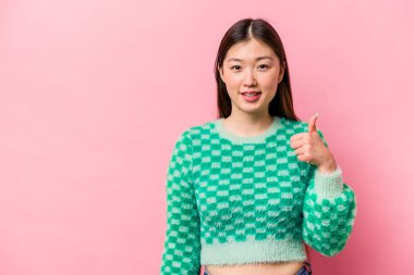 Genç Çinli bir kadın pembe arka planda izole edilmiş gülümsüyor ve başparmağını kaldırıyor.