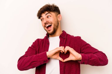 Beyaz arka planda izole edilmiş genç İspanyol adam gülümsüyor ve elleriyle kalp şeklini gösteriyor..