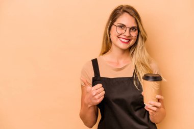 Restoran garsonu Kafkasyalı kadın elinde bej arka planda izole edilmiş kahve tutuyor gülümsüyor ve başparmağını kaldırıyor.