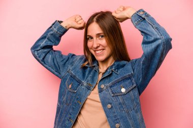 Pembe arka planda soyutlanmış genç beyaz bir kadın özel bir günü kutluyor, atlıyor ve enerjiyle kollarını kaldırıyor..