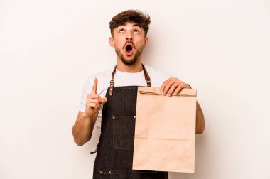 Beyaz arka planda izole edilmiş, ağzı açık bir şekilde duran bir paket taşıyan genç İspanyol tezgahtar..