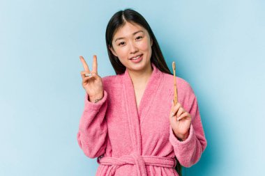 Bornoz giymiş genç Asyalı bir kadın pembe arka planda izole edilmiş dişlerini fırçalıyor neşeli ve tasasız parmaklarıyla barış sembolü gösteriyor..