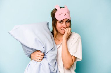 Mavi arka planda izole edilmiş pijama giyen genç beyaz kadın tırnaklarını yiyor, gergin ve çok endişeli..