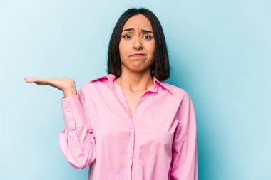 Latin kökenli genç bir kadın, mavi arka planda izole edilmiş. Sorgulama sırasında omuz silkiyor ve şüphe duyuyor..