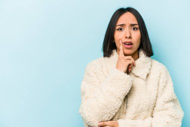 Mavi arka planda izole edilmiş genç İspanyol kadın şüpheli ve şüpheci bir ifadeyle yana bakıyor..
