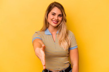 Sarı arka planda izole edilmiş genç beyaz kadın selamlaşma jesti olarak kameraya elini uzatıyor..