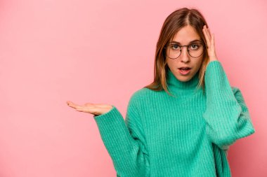 Pembe arka planda izole edilmiş genç beyaz kadın, avuç içi fotokopi alanını elinde tutmaktan etkilenmiş..