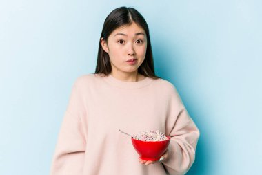 Genç Asyalı bir kadın mavi arka planda izole edilmiş bir kase mısır gevreği tutarken omuzlarını silker ve gözleri açık kalır..