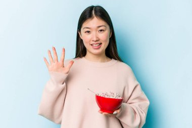 Mavi arka planda bir kase mısır gevreği tutan genç Asyalı kadın gülümseyerek beş numarayı parmaklarıyla gösteriyor..