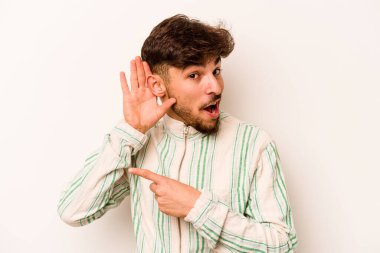 Latin kökenli genç bir adam beyaz arka planda bir dedikoduyu dinlemeye çalışıyor..