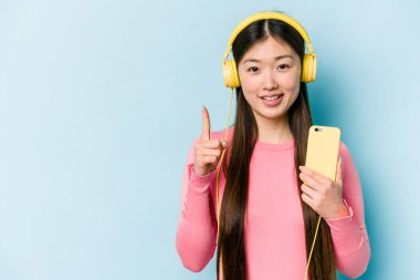Genç Asyalı müzik dinleyen bir kadın mavi arka planda bir numarayı parmağıyla gösteriyor..