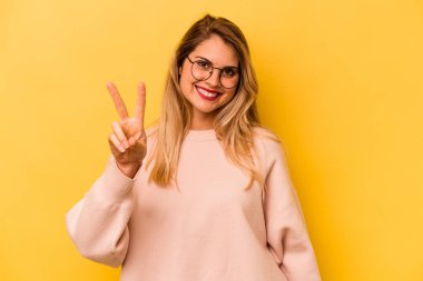 Sarı arka planda izole edilmiş genç beyaz kadın neşeli ve kaygısız parmaklarıyla barış sembolü gösteriyor..