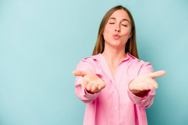 Mavi arka planda izole edilmiş genç, beyaz bir kadın. Dudaklarını kıvırıyor ve avuç içlerine hava öpücüğü yolluyor..