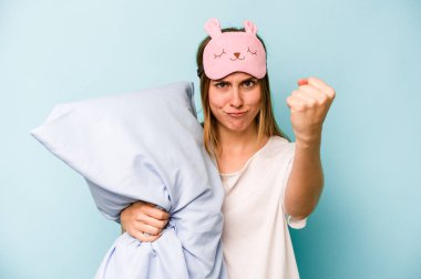 Mavi arka planda izole edilmiş pijama giyen genç beyaz kadın kameraya yumruk gösteriyor, agresif yüz ifadesi..