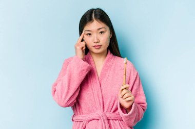 Bornoz giymiş genç Asyalı bir kadın pembe arka planda izole edilmiş dişlerini fırçalıyor. Şakağı parmağıyla işaret ediyor, düşünüyor, bir göreve odaklanıyor..