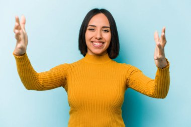 Genç İspanyol kadın mavi arka planda izole edilmiş zaferi ya da başarıyı kutluyor, şaşırmış ve şok olmuş..