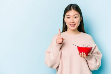 Genç Asyalı bir kadın elinde bir kase mısır gevreği tutarken mavi arka planda bir numarayı parmağıyla gösteriyor..