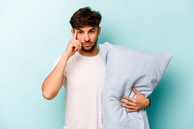 Mavi arka planda izole edilmiş pijama giyen genç İspanyol bir adam şakağı parmağıyla işaret ediyor, düşünüyor ve bir göreve odaklanıyor..