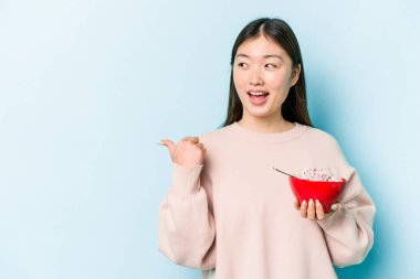 Genç Asyalı bir kadın elinde bir kase mısır gevreği tutarken mavi arka plan noktalarından izole edilmiş başparmak uzakta, gülerek ve kaygısızca.