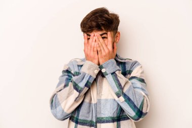 Beyaz arka planda izole edilmiş genç İspanyol adam kameraya parmaklarıyla göz kırpıyor, yüzünü kapatmaktan utanıyor..