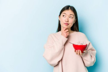 Genç Asyalı bir kadın elinde bir kase mısır gevreği tutarken mavi arka planda izole edilmiş şüpheli ve şüpheci bir ifadeyle yana bakıyor..