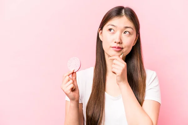 年轻的亚洲女人 手里拿着用粉红背景隔开的面部海绵 侧视着对方 脸上带着怀疑和怀疑的表情 — 图库照片