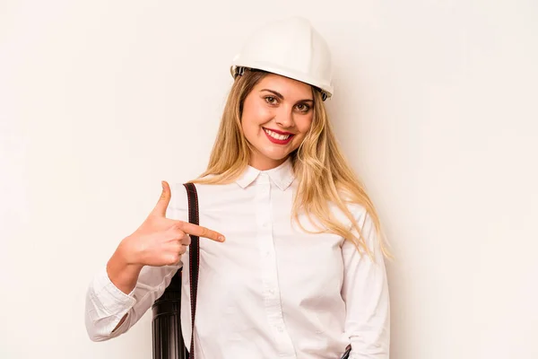 Young architect woman with helmet and holding blueprints isolated on white background person pointing by hand to a shirt copy space, proud and confident