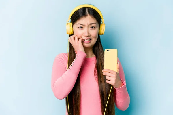 Jovem Mulher Asiática Ouvindo Música Isolada Fundo Azul Mordendo Unhas — Fotografia de Stock
